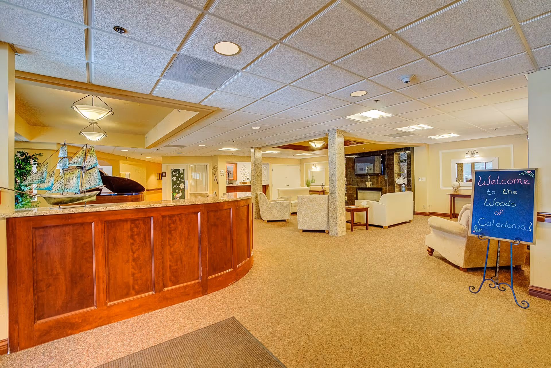 Interior view of a senior living facility lobby area with a wooden reception desk on the left, a decorative model ship on the desk, comfortable seating with armchairs and sofas, a fireplace with a mounted TV above it, and a chalkboard sign on the right that reads 'Welcome to the Woods of Caledonia!'