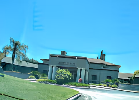 Exterior view of a single-story building with a sloped roof, surrounded by green grass and some bushes, under a clear blue sky.