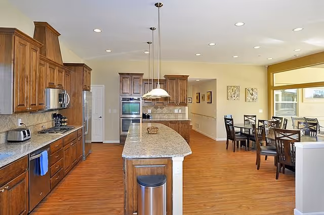 A spacious kitchen and dining area with wooden cabinets, granite countertops, and hardwood floors. The kitchen features a large island with a trash bin underneath, stainless steel appliances including a refrigerator, microwave, and oven. The dining area has multiple dark wooden tables and chairs near large windows letting in natural light.