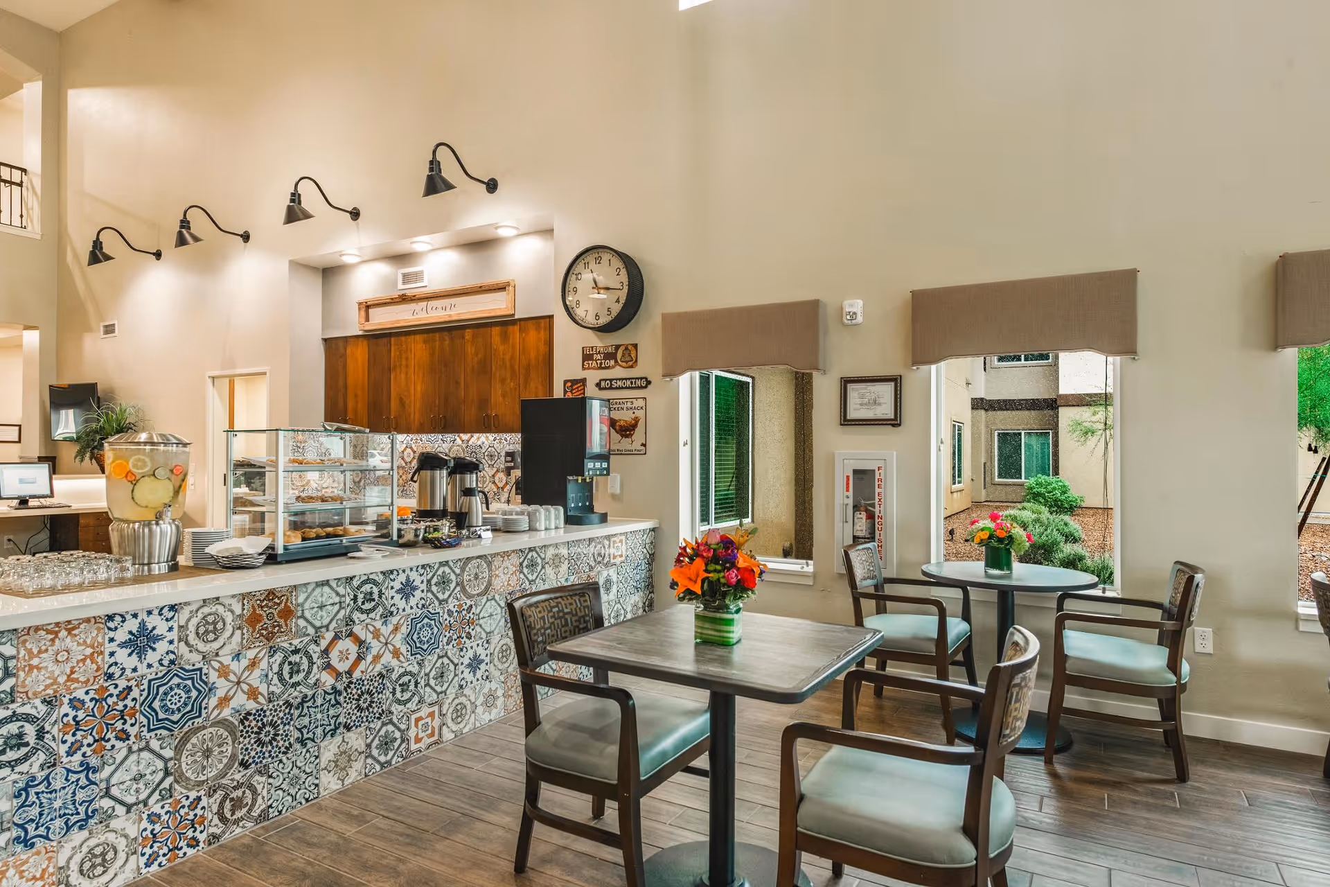 A bright and inviting dining area in a senior living facility featuring a counter with colorful patterned tiles, a beverage dispenser, coffee station, and two small tables with chairs. The walls are light-colored with windows showing greenery outside, and there are wall-mounted lights and a clock above the counter.