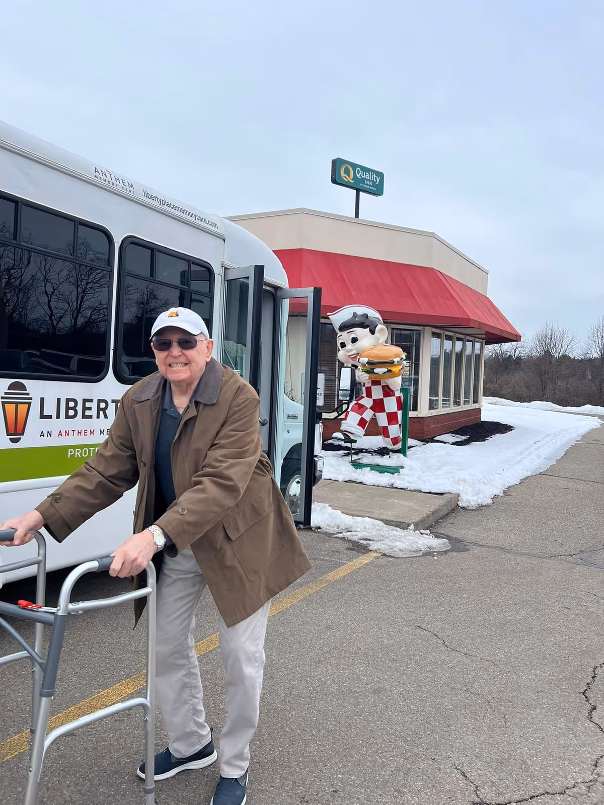 An elderly man using a walker is smiling and standing outside near a Liberty Place Memory Care shuttle bus. Behind him is a building with a red awning and a large statue of a character holding a hamburger. Snow is visible on the ground around the building.