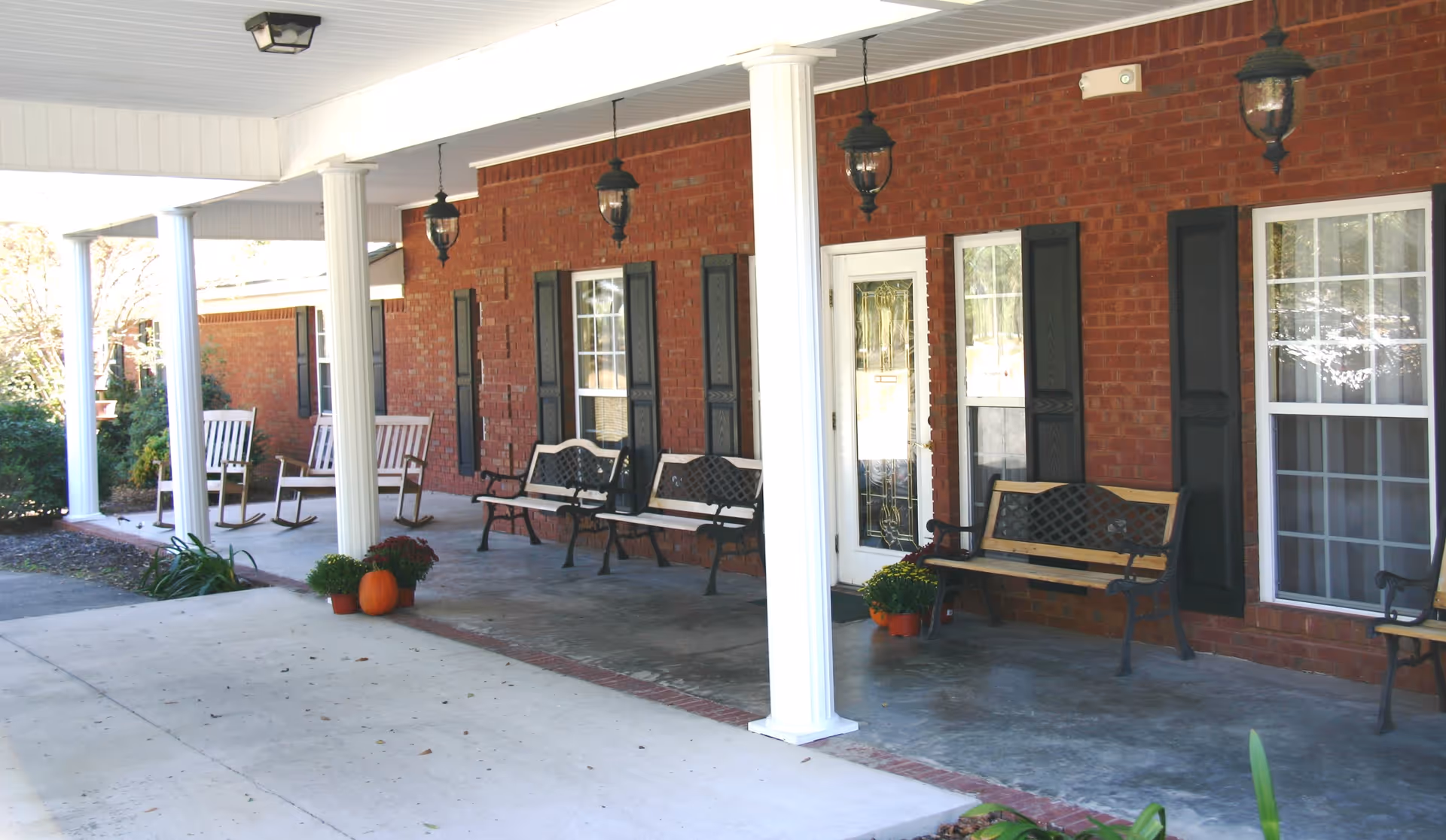 Covered porch area of a brick building with white columns, black shutters on windows, several benches and rocking chairs, hanging lantern-style lights, and potted plants including a pumpkin and flowers.