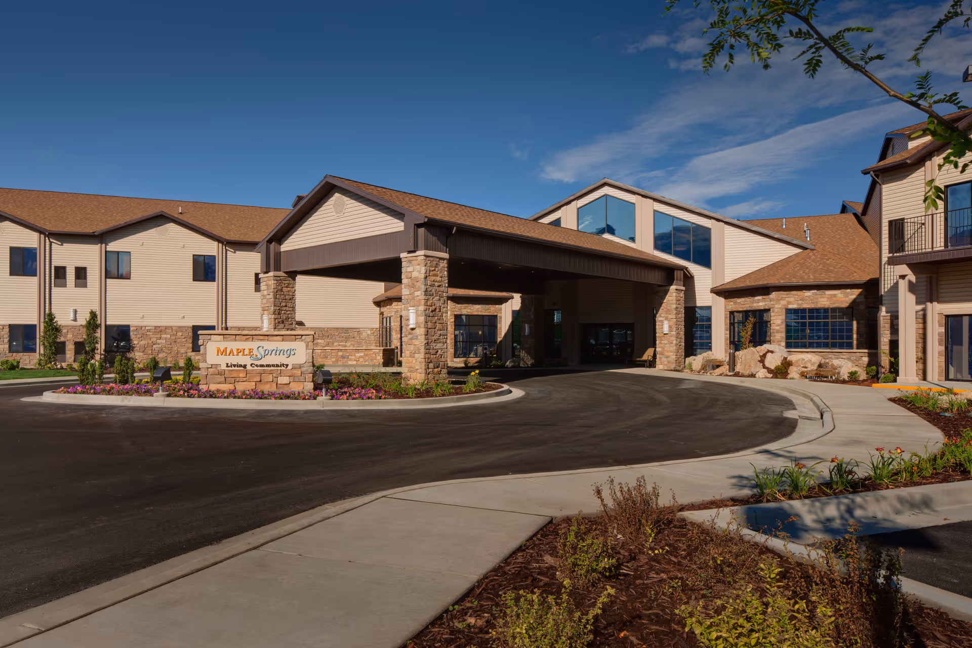 Exterior view of Maple Springs of Brigham City senior living community building with a covered entrance, stone and beige siding, landscaped flower beds, and a clear blue sky.