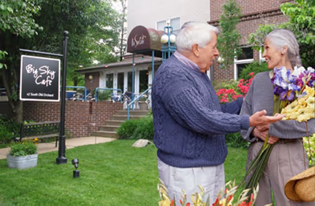 An elderly man and woman stand outside near a garden area in front of a building. The man is wearing a blue sweater and white pants, and the woman is holding a bouquet of flowers. Behind them is a sign that reads 'Big Sky Cafe' and a building entrance with steps and a canopy.