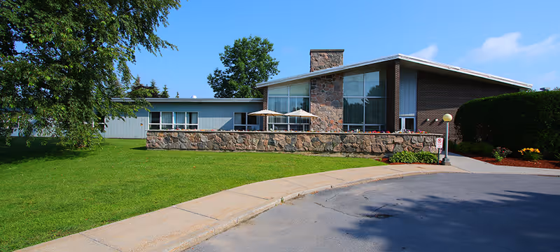 Exterior view of a single-story building with a stone facade and large windows, surrounded by green grass and trees under a clear blue sky. There is a curved driveway and a sidewalk leading to the entrance.