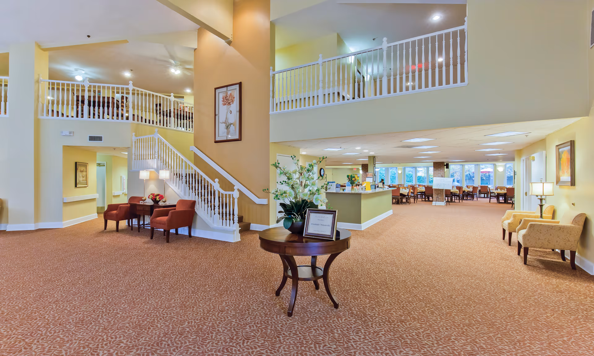 Spacious senior living lobby with a central round table, staircase, seating areas, reception desk, and an upper balcony railing.