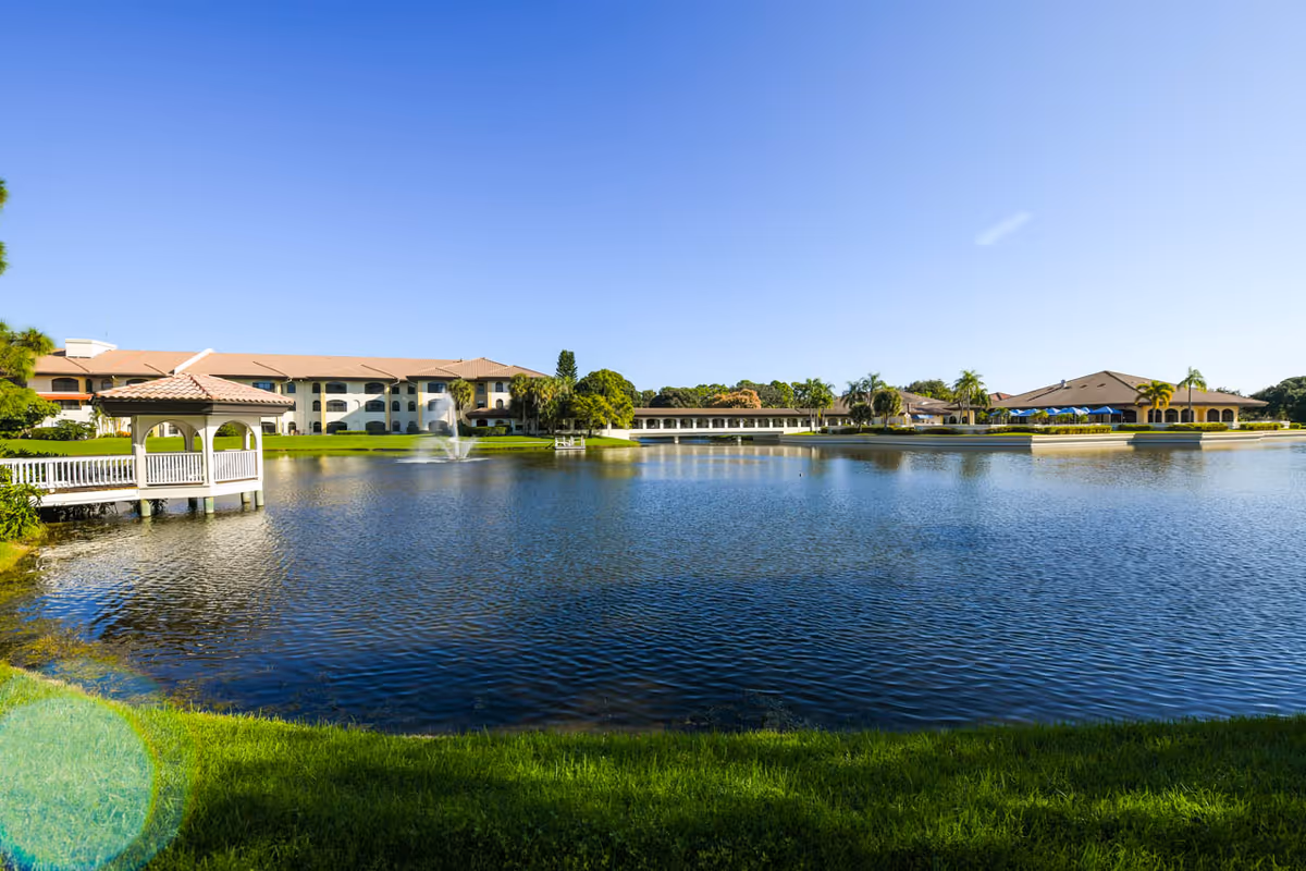 A serene outdoor view of The Barclay at Sarasota facility featuring a large pond with a water fountain in the center, surrounded by green grass and palm trees. The buildings of the facility are visible in the background under a clear blue sky, with a small gazebo on the left side extending over the water.