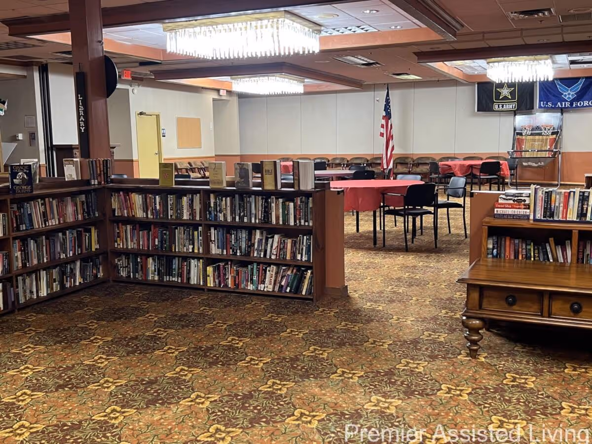 A library area with low bookshelves in the foreground and a community room with round tables, chairs, and an American flag in the background.
