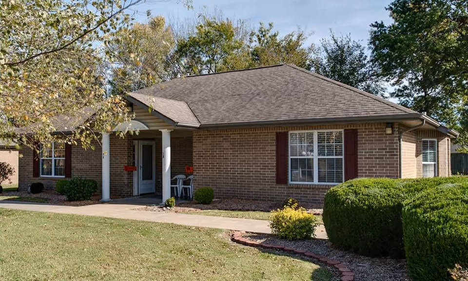 Single-story brick building with a gabled roof, white columns at the entrance, and windows with dark shutters. The building is surrounded by green bushes, a small tree, and a lawn with a paved walkway leading to the entrance. The sky is clear with some clouds.