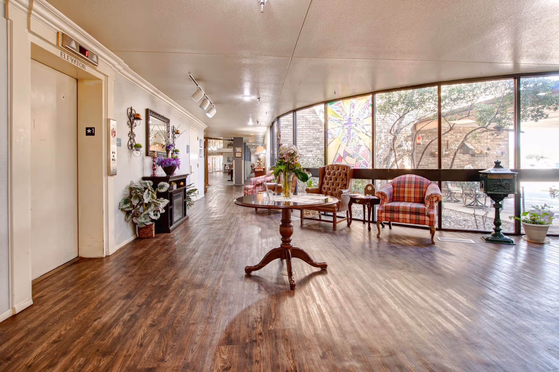 A bright and spacious hallway in a senior living facility with wooden flooring, an elevator on the left, and a seating area on the right featuring a round wooden table with a flower vase, a brown leather armchair, and a plaid upholstered armchair. Large windows let in natural light and show an outdoor garden area with trees and a colorful stained glass panel.