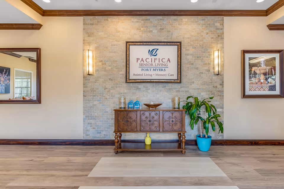 Interior view of a senior living facility lobby area featuring a wooden console table with decorative items, a potted plant in a blue pot, two wall sconces, a framed sign that reads 'Pacifica Senior Living Fort Myers Assisted Living Memory Care', and two framed pictures on either side of the stone accent wall.