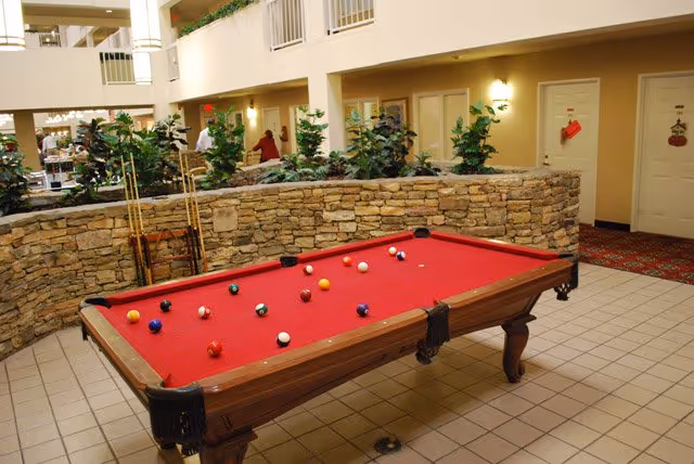 Indoor common area in a retirement community with a red-felt pool table in front of a low stone planter wall and surrounding doors and balconies.