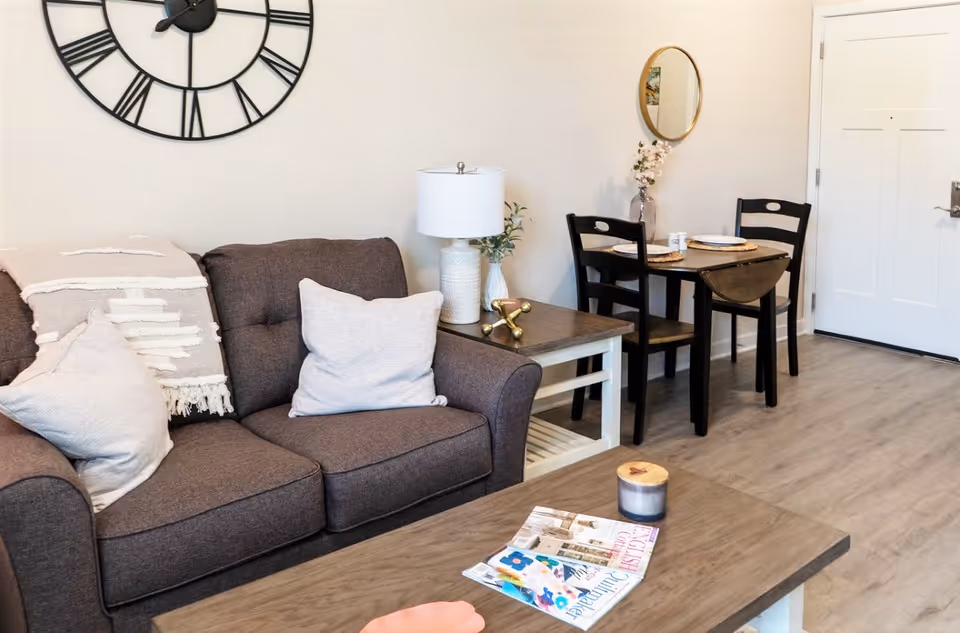 A cozy living area featuring a dark gray sofa with light-colored pillows and a throw blanket, a wooden coffee table with a candle and magazines, a side table with a white lamp and decorative items, a small dining table set with two black chairs, a round wall mirror, and a large wall clock with Roman numerals. The room has light-colored walls and wood flooring.