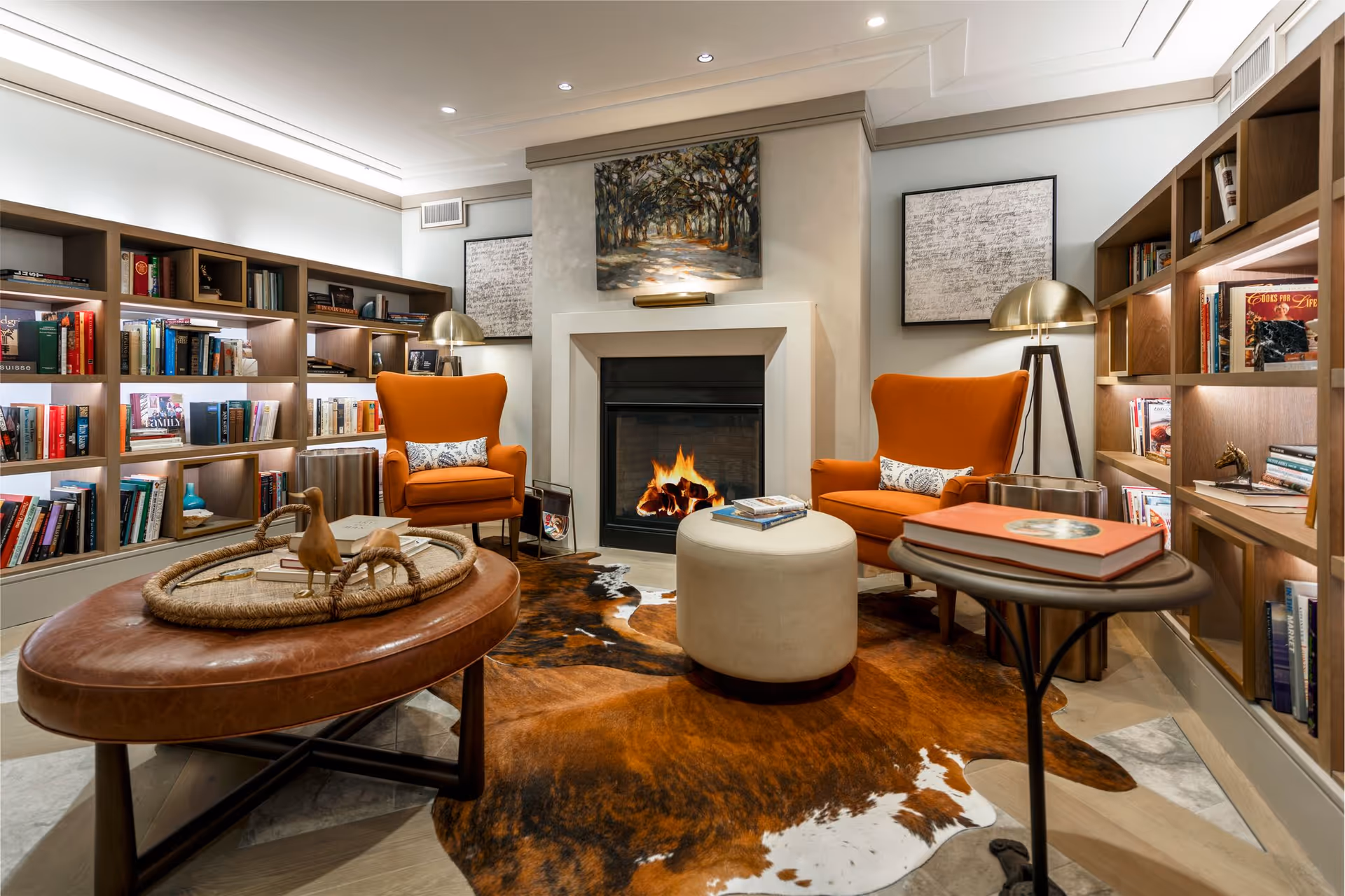 Cozy seating area with two orange armchairs facing a fireplace, built-in bookshelves, a leather ottoman and a cowhide rug.