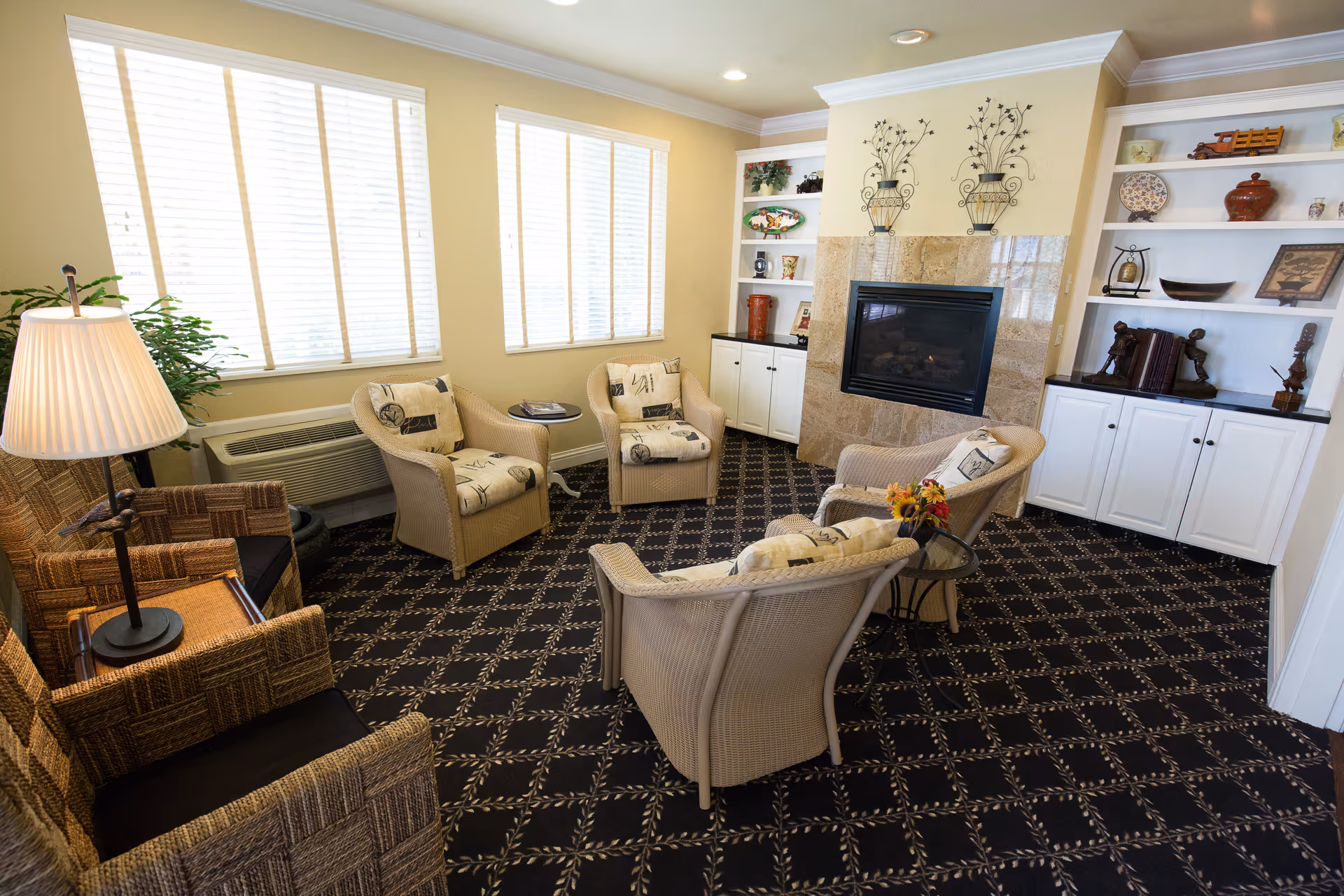 A cozy living room area with five cushioned wicker chairs arranged around a small glass-top table with a flower arrangement. The room features a patterned carpet, a fireplace with a tiled surround, built-in white shelves and cabinets filled with decorative items, and large windows with white blinds letting in natural light.