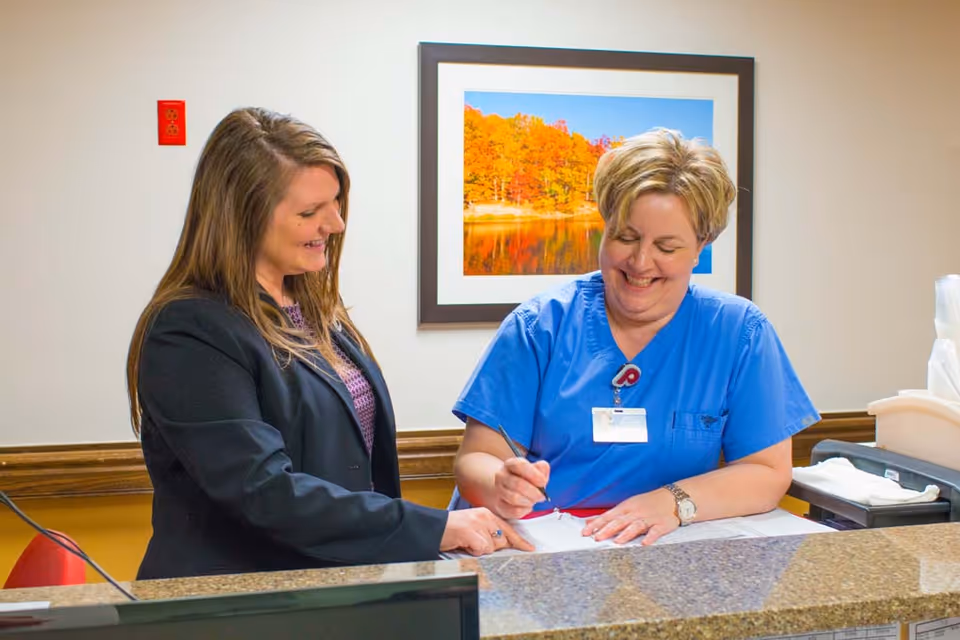 A woman in a black blazer and a woman in blue medical scrubs are standing at a reception desk. The woman in scrubs is smiling and writing on a document while the other woman points at the paper. Behind them is a framed picture of a lake with autumn trees.