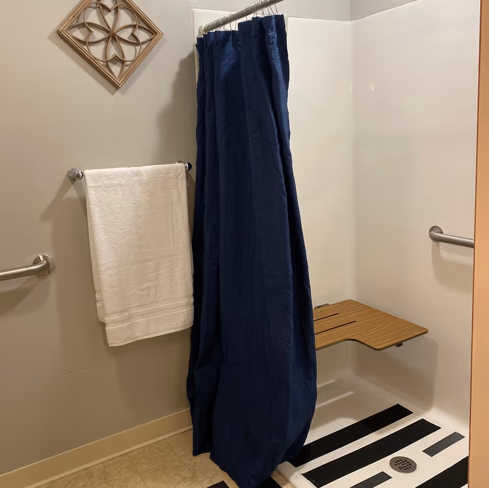 A bathroom shower area with a navy blue shower curtain, a white towel hanging on a towel rack, a wooden fold-down shower seat, and grab bars on the walls. The floor has black and white striped pattern near the drain.