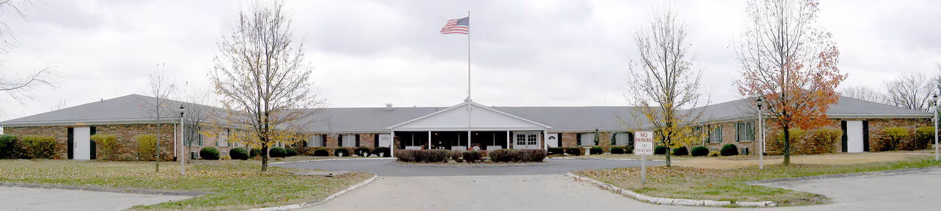 Single-story brick care facility with a central covered entrance, American flag on a flagpole, and a semicircular driveway.