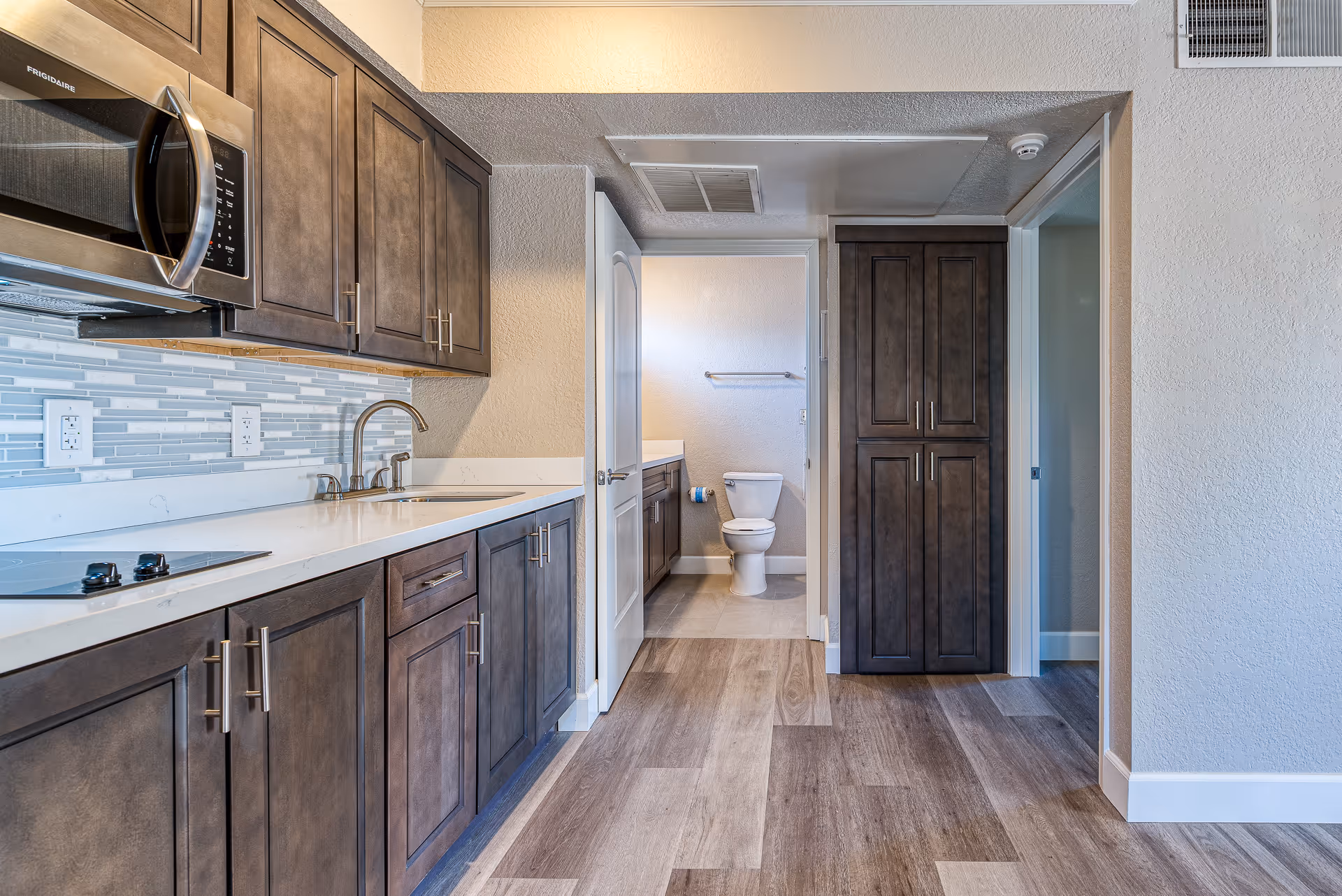 Modern kitchen area with dark wood cabinets, a white countertop, a built-in microwave, and a stovetop. In the background, there is a bathroom with a visible toilet and a vanity with a sink. The flooring is wood-style laminate, and the walls are painted light beige.