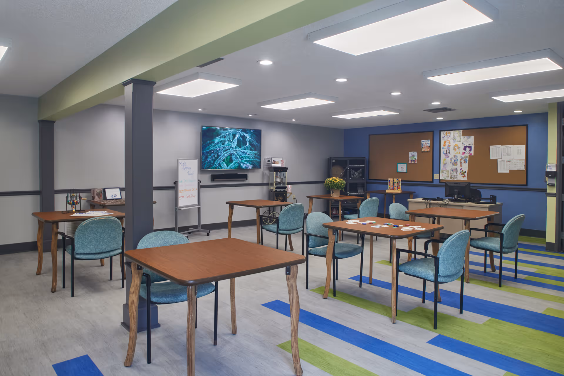 Bright multipurpose activity room with several wooden tables and teal chairs, a wall-mounted TV, bulletin boards, and colorful blue-and-green striped flooring.