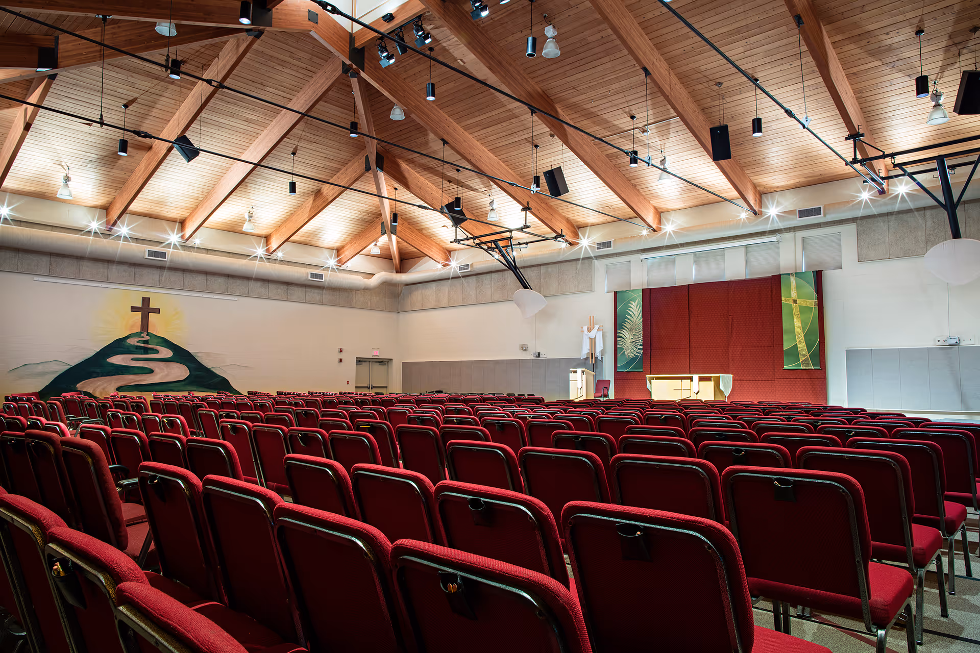 Interior view of a large chapel or worship hall with rows of red cushioned chairs facing a stage with an altar. The ceiling features exposed wooden beams and multiple lights. A mural of a cross on a hill with a winding path is painted on the left wall.