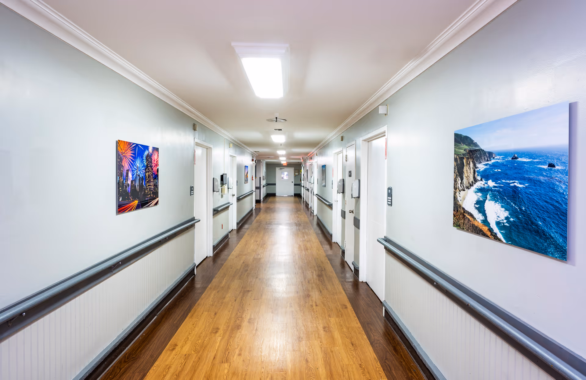 A long, well-lit hallway in a nursing center with wooden flooring and light gray walls. The hallway has handrails on both sides and several closed white doors along the walls. Colorful framed pictures, including one of fireworks and another of a coastal scene, hang on the walls.