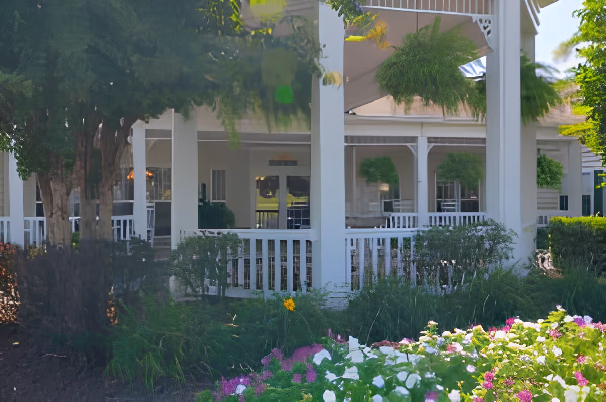 A white porch with railings and hanging green plants, surrounded by lush greenery and colorful flowers in the foreground, attached to a building.