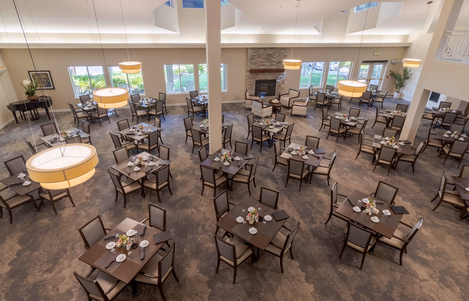 Spacious dining room in a retirement community with multiple square tables set with plates, cups, and napkins. The room features large windows letting in natural light, a stone fireplace with seating around it, hanging pendant lights, and a piano in the corner.