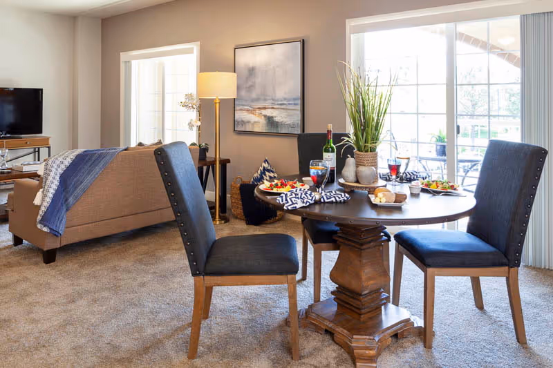 Bright open living and dining area with a round wooden table set for a meal, upholstered chairs, a sofa, and large sliding glass doors.