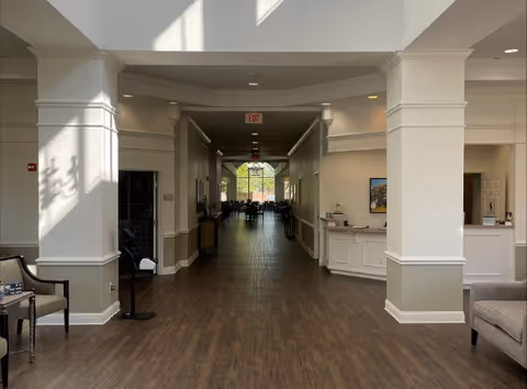 Interior view of a senior living facility hallway with wood flooring, white walls, and columns. There are chairs and a small table on the left, a reception desk on the right, and a dining area visible at the far end of the hallway with large windows letting in natural light.