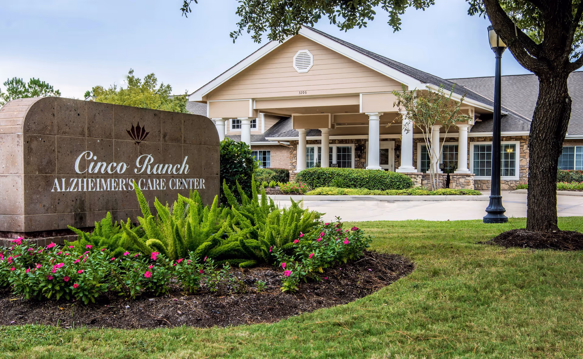 Exterior view of Cinco Ranch Alzheimer's Care Center showing the main entrance with white columns and a landscaped garden with green plants and pink flowers in front of a large stone sign displaying the facility's name.
