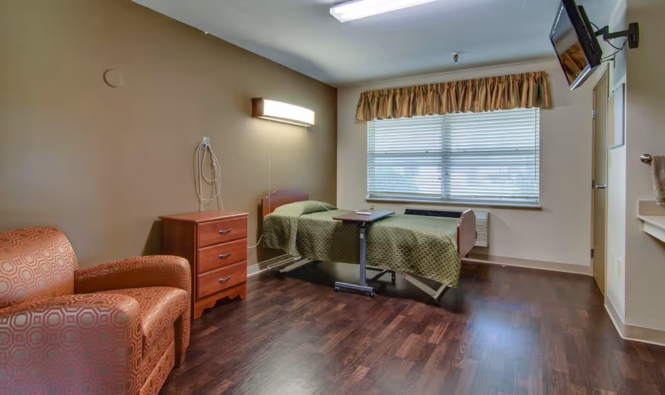 A senior living facility bedroom with a single bed covered in a green quilt, a wooden nightstand with three drawers, a patterned armchair, a wall-mounted light above the bed, a window with blinds and a valance, a wall-mounted TV, and a small overbed table on wheels. The room has wood flooring and neutral-colored walls.