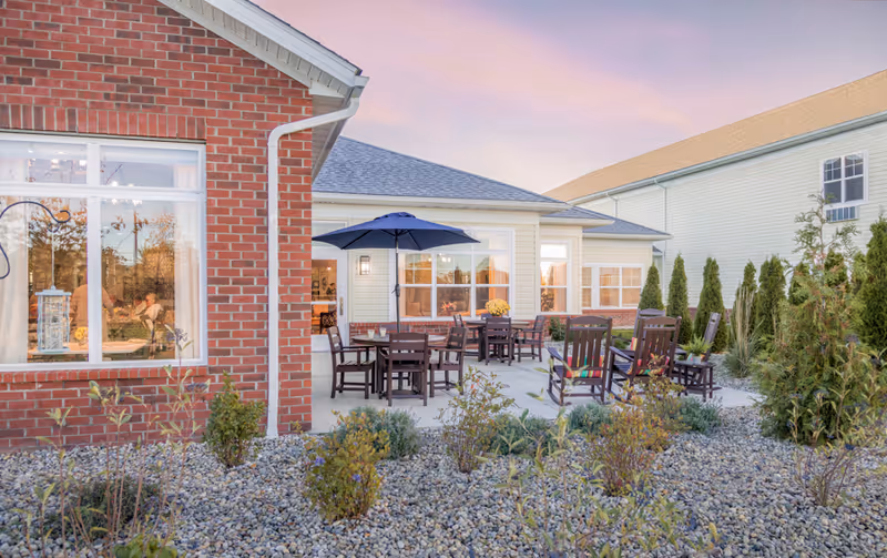 Outdoor patio area at Randall Residence of West Milton with several wooden tables and chairs, some under a large blue umbrella, surrounded by landscaping with small bushes and rocks. The building exterior features brick and light-colored siding with multiple windows.