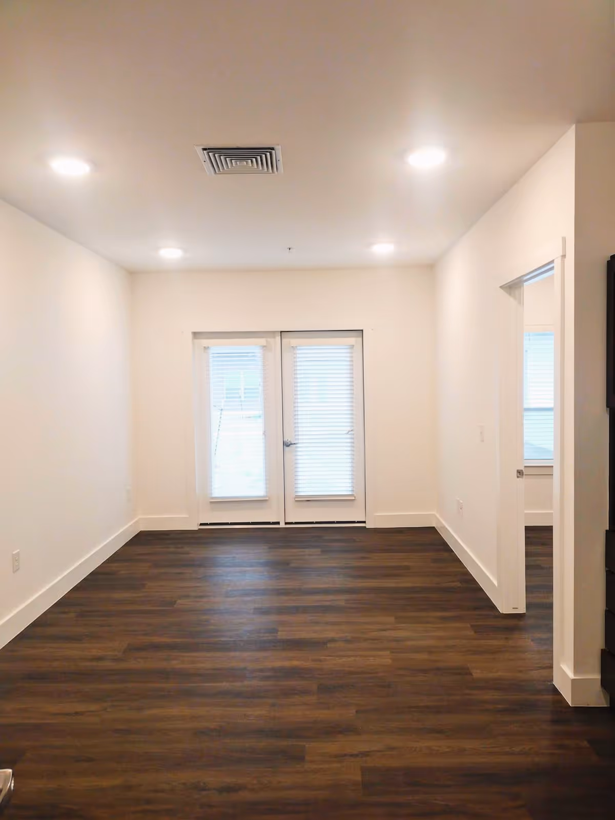 Empty room with dark wood flooring, white walls, and ceiling lights. There are double glass doors with blinds at the far end and an open doorway on the right side leading to another room.