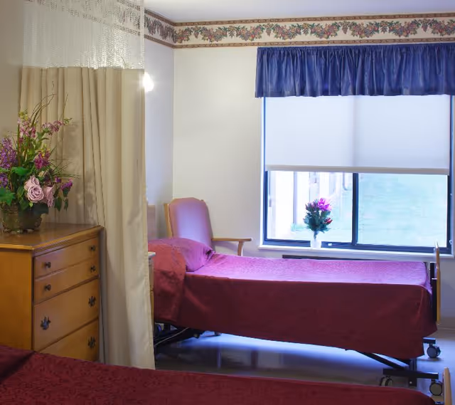 A senior living bedroom with an adjustable bed covered in a maroon bedspread near a window, a wooden dresser with a floral arrangement, a chair, and a privacy curtain.