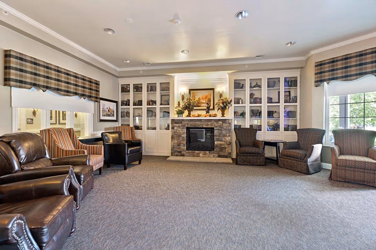 Spacious cozy living room with a stone fireplace flanked by built-in white bookshelves, multiple armchairs and plaid valances on the windows.