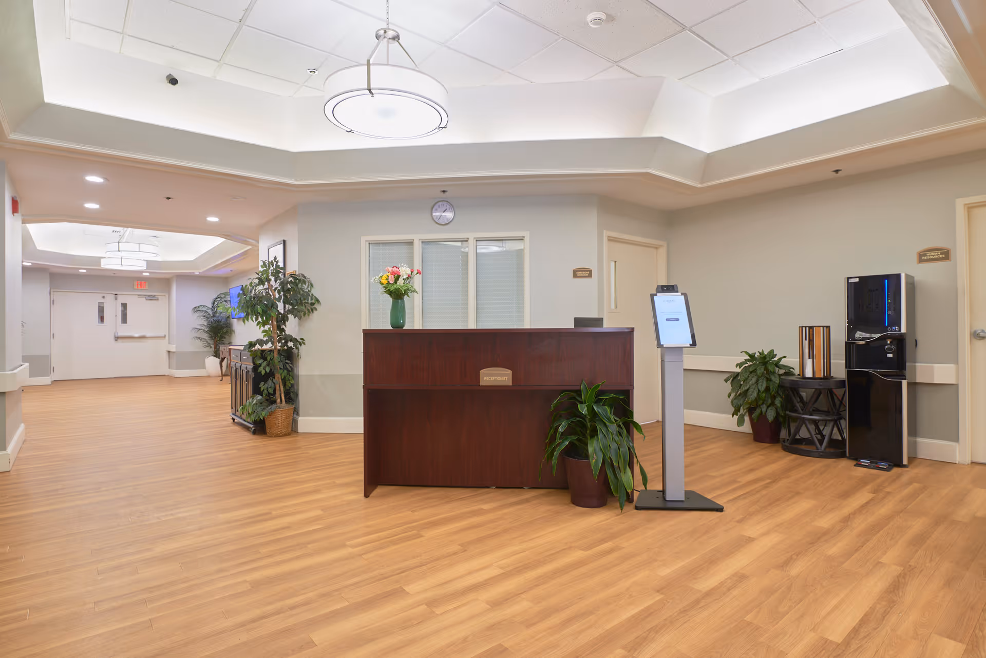 Reception desk in a well-lit senior living facility lobby with wood floors, plants, a tablet kiosk, and a water cooler.
