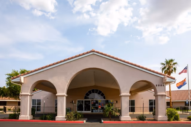 Front exterior view of South Mountain Post Acute building with a covered entrance supported by columns, a tiled roof, and flags on flagpoles to the right under a partly cloudy sky.