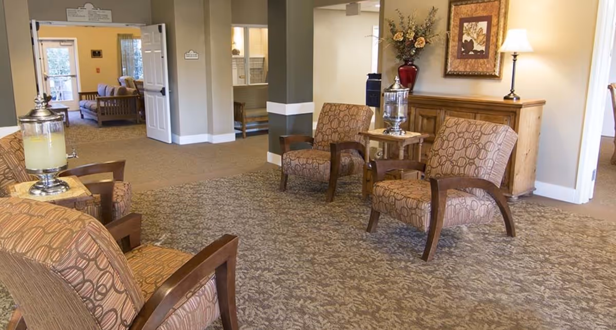 A cozy seating area in a senior living facility with four patterned armchairs arranged around two small wooden tables, each holding a beverage dispenser. The room has beige walls, a patterned carpet, a wooden cabinet with a lamp and floral arrangement, and an open doorway leading to another room with additional seating.