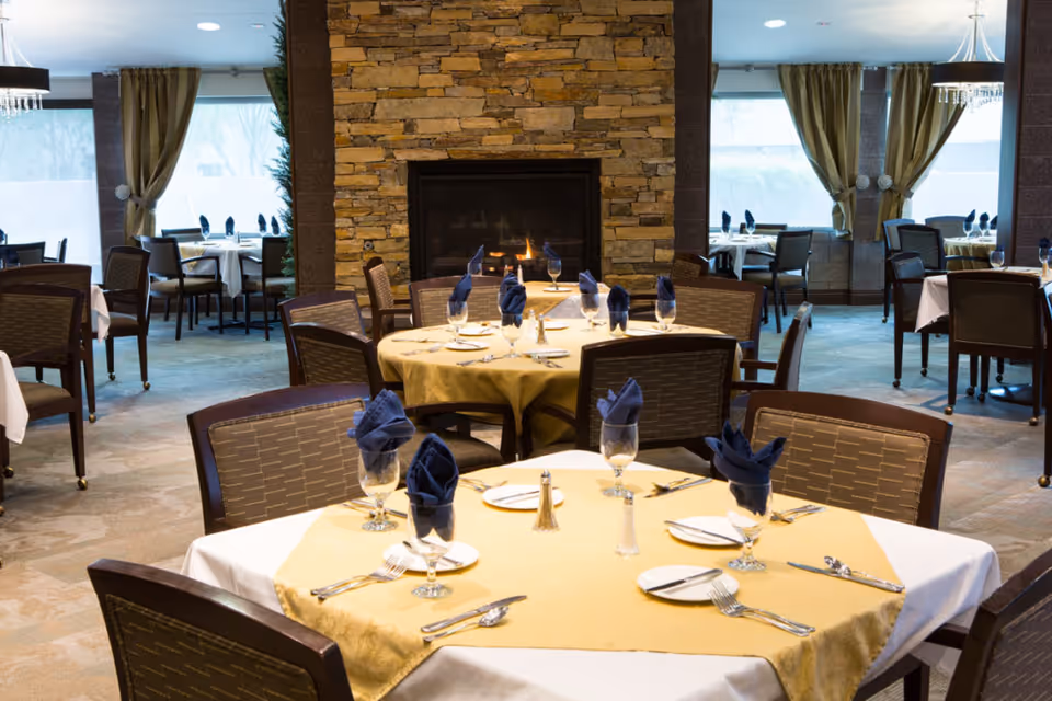 Elegant dining room with round tables set with yellow tablecloths, folded blue napkins, glassware and a central stone fireplace.