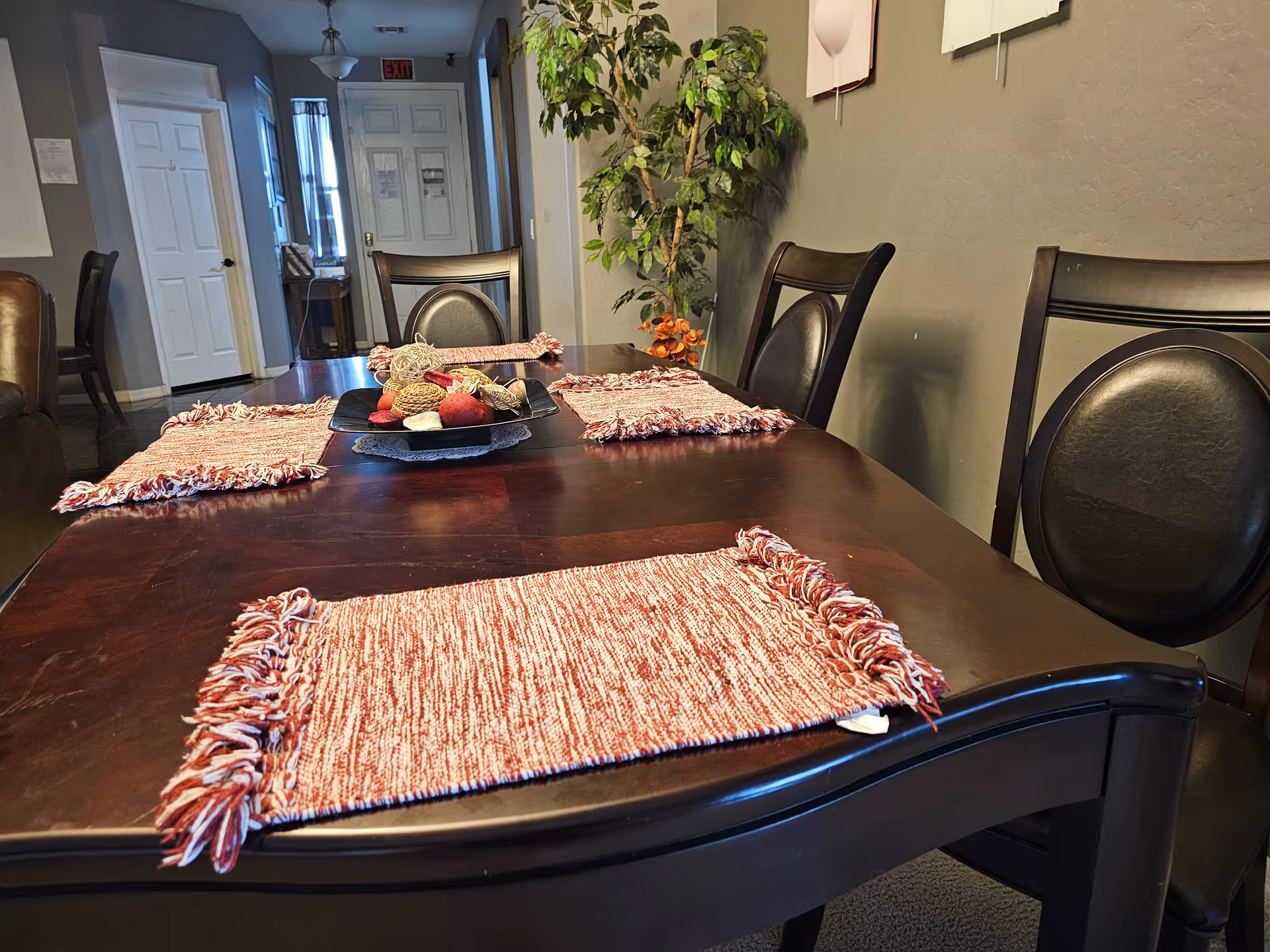 A dining room with a dark wooden table set with four red and white woven placemats. The table has a decorative centerpiece with various dried natural elements. Surrounding the table are dark wooden chairs with black cushioned seats and backs. In the background, there is a hallway with white doors and an exit sign above one door. A tall green potted plant is placed against a beige wall.