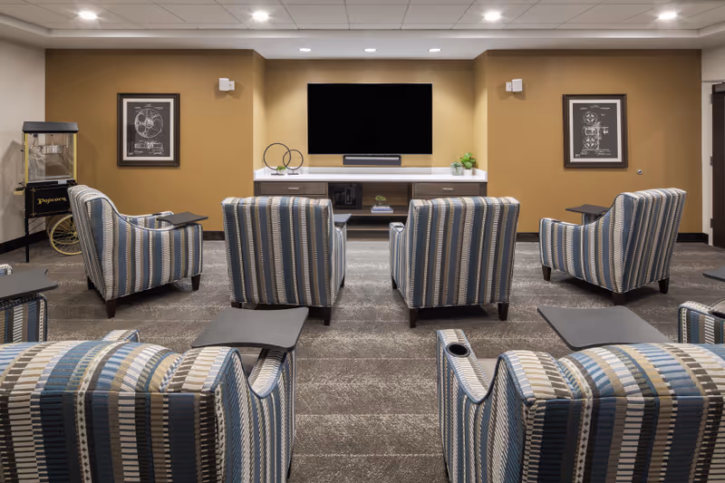 A cozy media room with six striped armchairs arranged in two rows facing a large flat-screen TV mounted on a beige wall. The room features a popcorn machine on the left side, two framed pictures on the walls, and a console table beneath the TV with decorative items and books. The carpet is patterned in neutral tones, and the ceiling has recessed lighting.