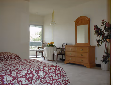 A bedroom in Gardena Retirement Center featuring a bed with a red and white floral patterned bedspread, a wooden dresser with a mirror, two chairs with a small round table covered with a white tablecloth and a flower arrangement, a hanging light fixture, and a large window with a view of greenery outside.
