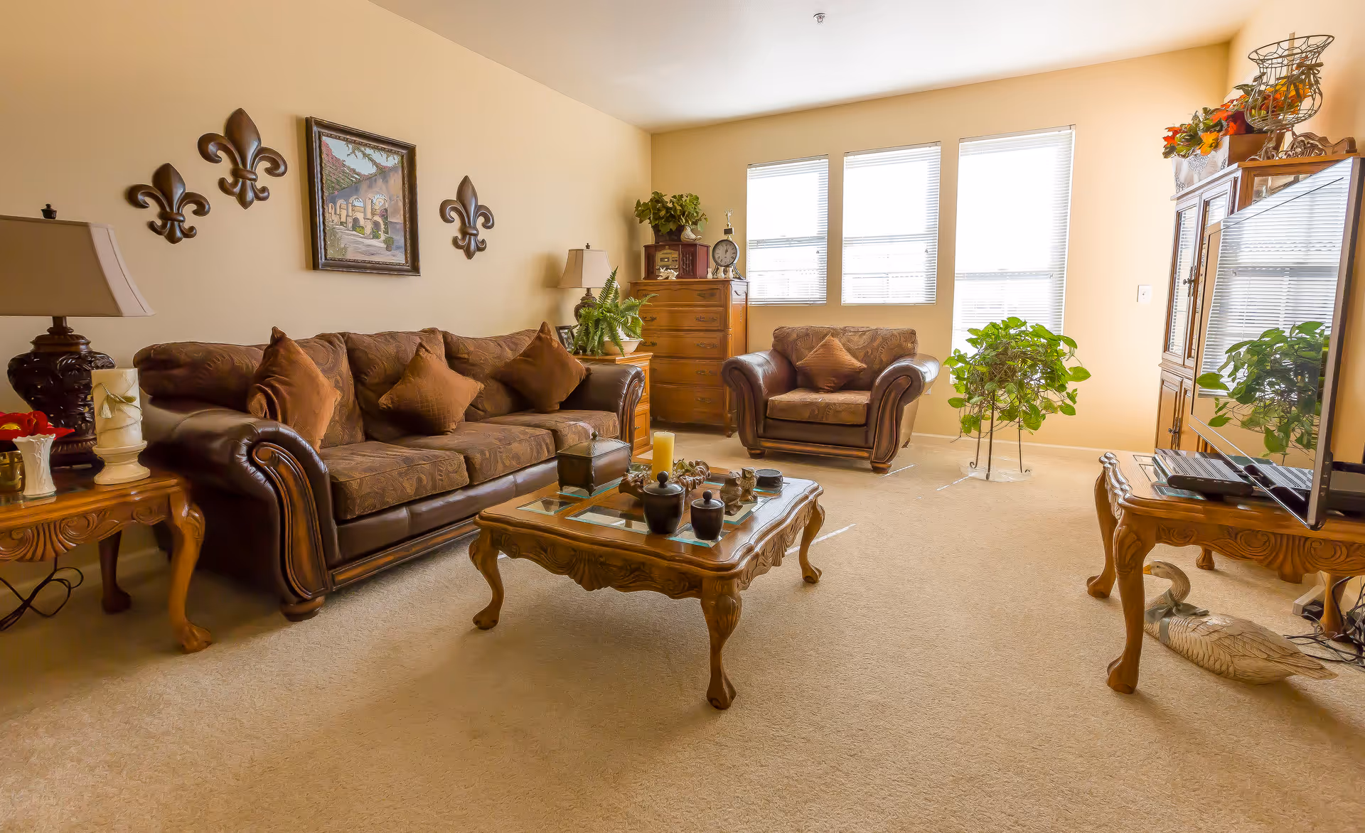 A cozy living room with a brown leather sofa and matching armchair, both adorned with brown cushions. A wooden coffee table with decorative items sits in the center on a beige carpet. The room has beige walls with three fleur-de-lis wall decorations and a framed painting. There are two wooden side tables with lamps and plants, a wooden chest of drawers, and a large window letting in natural light. A flat-screen TV is placed on a wooden TV stand on the right side of the room.
