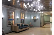 Interior view of a hallway or common area in a care facility featuring a striped sofa with blue cushions, three framed landscape paintings on the wall, a decorative round mirror, wall sconces, and ceiling chandeliers with multiple lights. The floor is light-colored, and the walls are painted in a soft green shade.