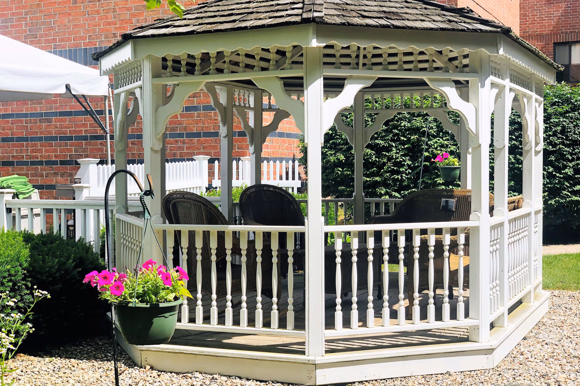 A white wooden gazebo with a shingled roof situated outdoors on a gravel area. Inside the gazebo are several wicker chairs arranged for seating. There are hanging flower pots with pink flowers on the outside of the gazebo. In the background, there is a brick building, green bushes, and a white picket fence.