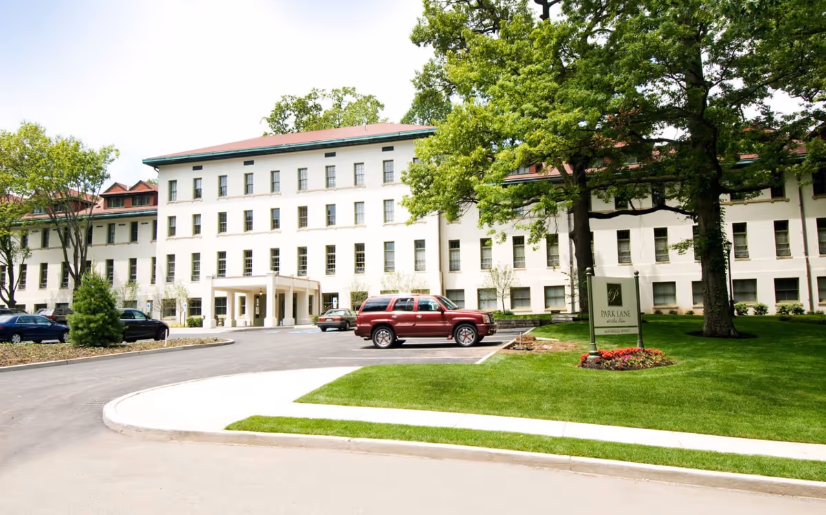 Exterior view of a large, white multi-story building with a red roof, surrounded by green trees and a well-maintained lawn. Several cars are parked in front of the building near the entrance. A sign on the lawn reads 'Park Lane at Sea View'.