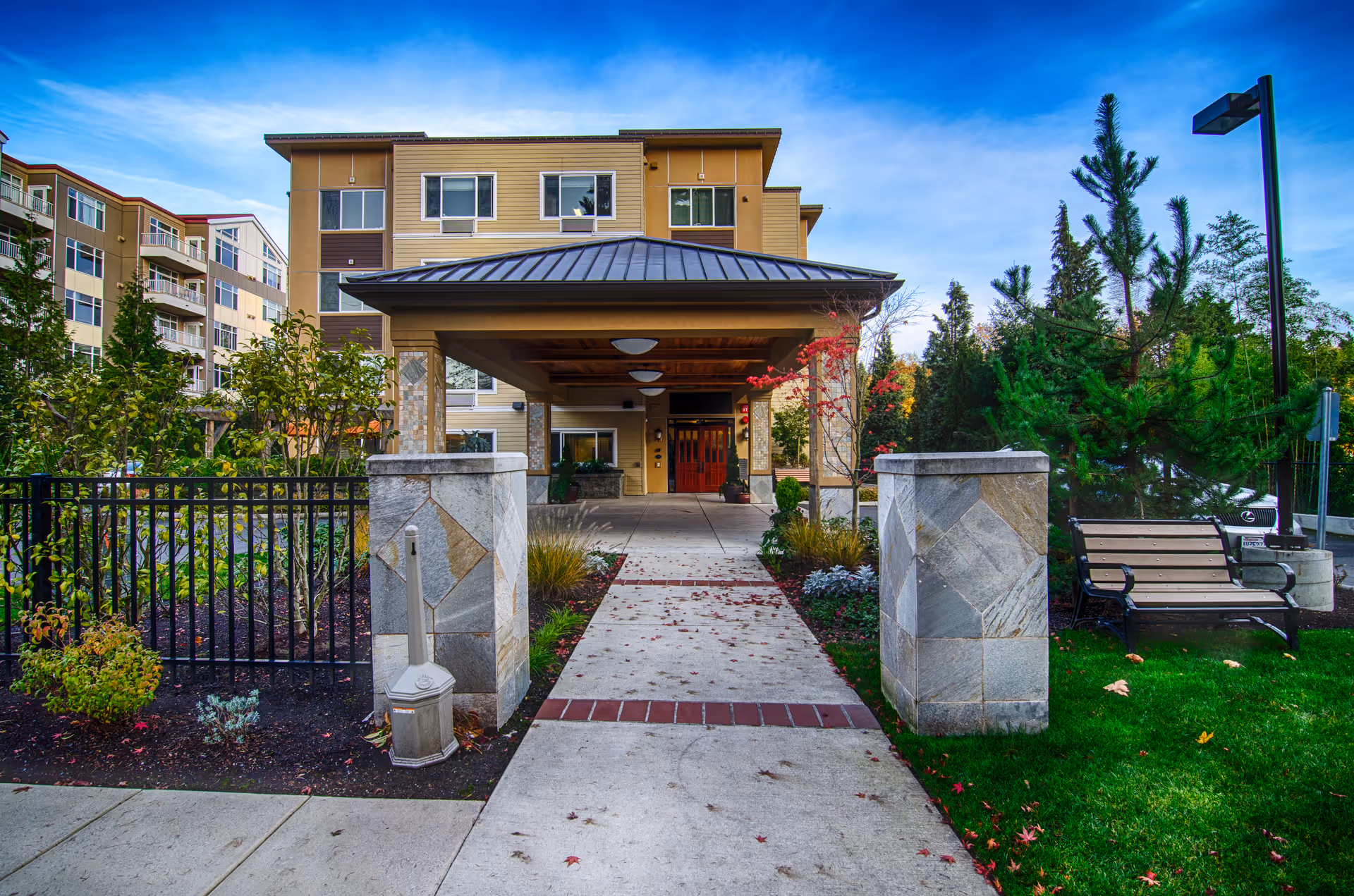 Entrance to a multi-story senior living facility with a covered drop-off area, surrounded by landscaped greenery, a bench, and a paved walkway leading to the front doors under a clear blue sky.