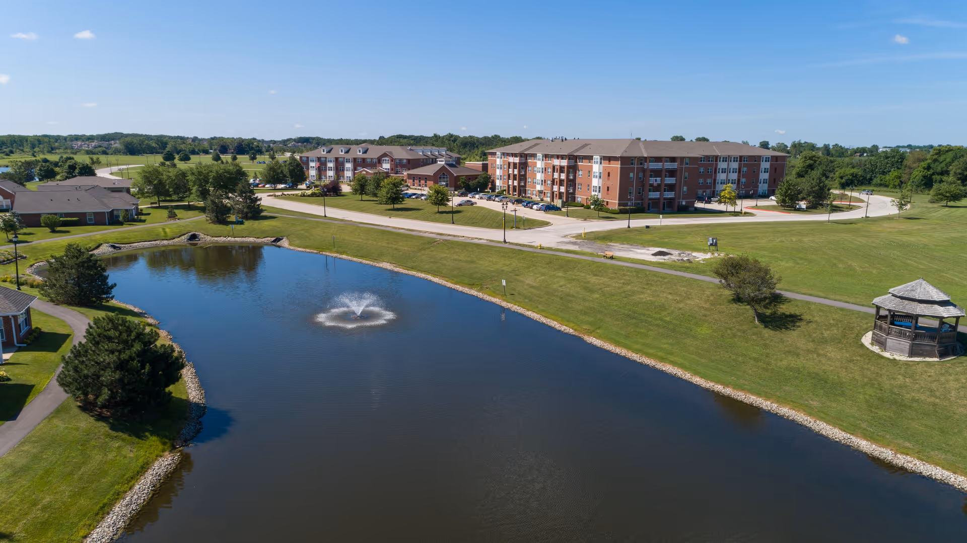 Aerial view of Marian Village senior living facility showing a large pond with a fountain in the center, surrounded by green lawns, trees, a gazebo, and multiple residential buildings under a clear blue sky.