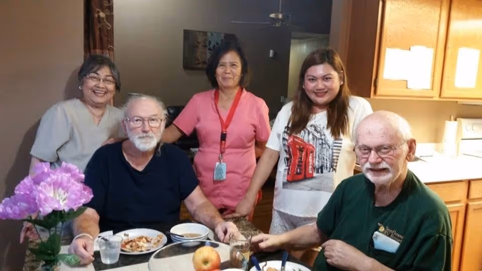 Two elderly men sitting at a dining table with plates of food and drinks, accompanied by three women standing behind them in a kitchen or dining area. There is a vase with purple flowers on the table and wooden cabinets in the background.